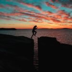 silhouette photo of a man jumps on to cliff near sea during sunset